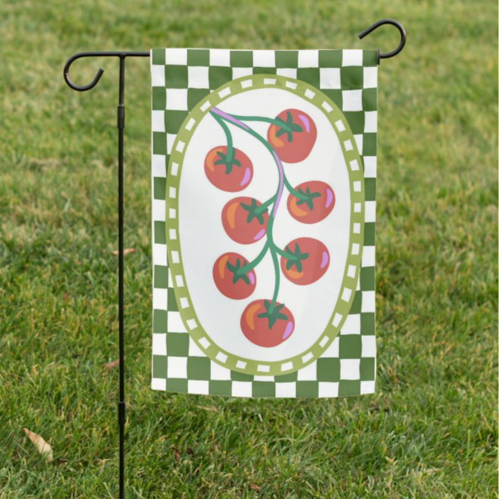 Garden flag with tomato design on a green and white checkered background, attached to a pole in grass.